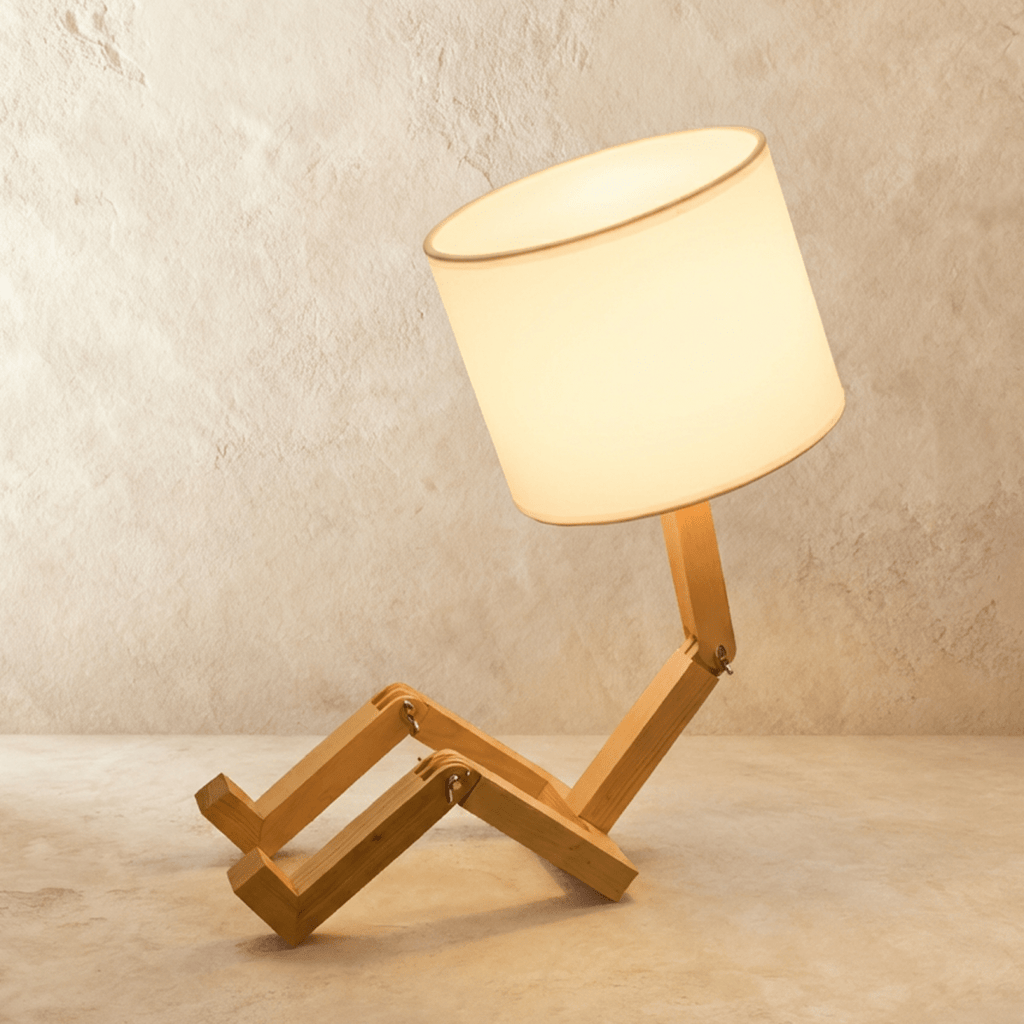 Clérachic wooden table lamp with a jointed, seated humanoid frame and a lit drum fabric shade, on a limewashed backdrop.