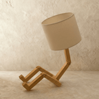 Clérachic wooden table lamp with jointed legs bent in a seated pose and a cream drum fabric shade, switched off.