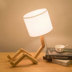 Clérachic wooden table lamp with a seated, jointed frame and white drum shade glowing on a wooden desk beside books and a cup.