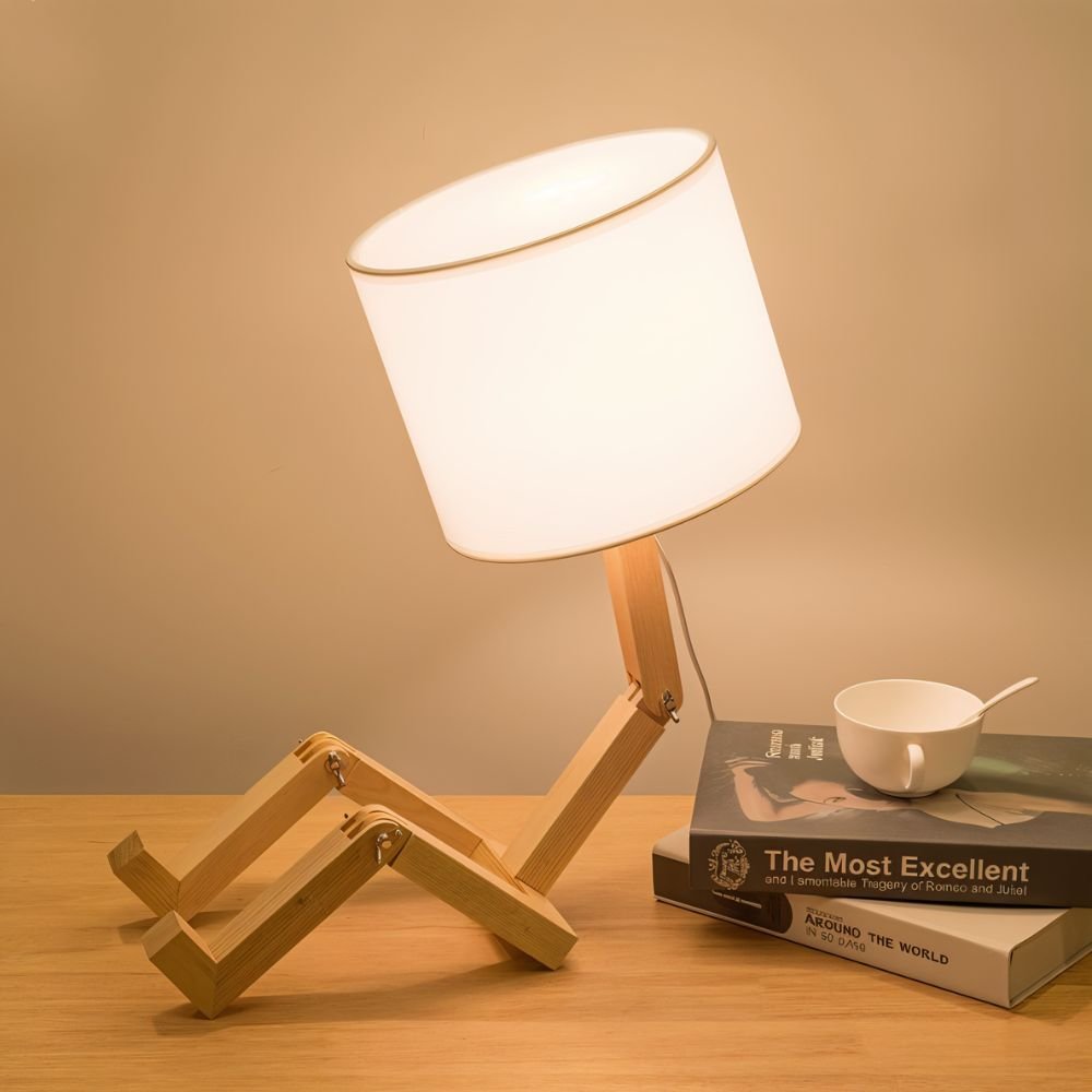 Clérachic wooden table lamp with a seated, jointed frame and white drum shade glowing on a wooden desk beside books and a cup.