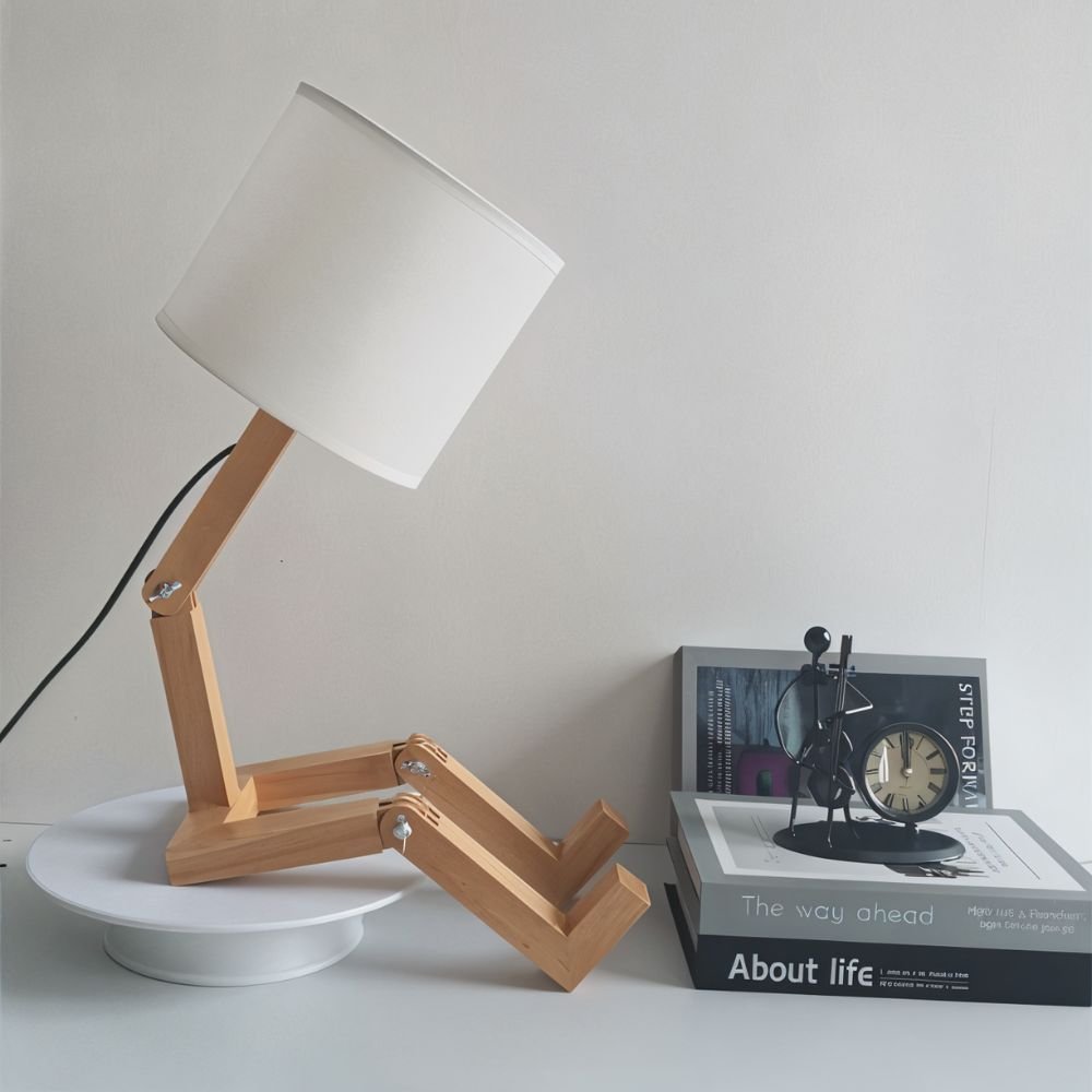 Clérachic seated wooden figure table lamp with a white fabric drum shade, shown unlit on a white turntable beside books and a clock.