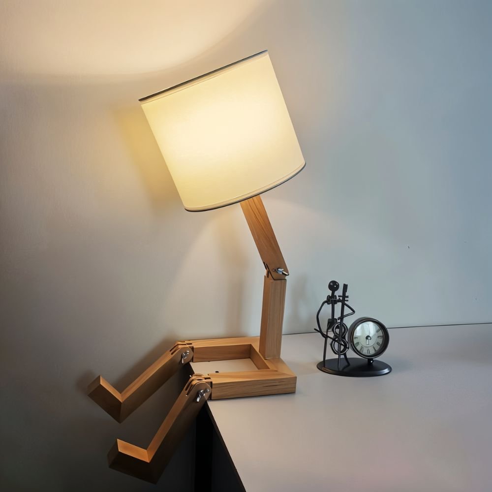 Clérachic wooden table lamp leaning on a desk edge with legs hanging over, its drum shade glowing beside a small musician clock.