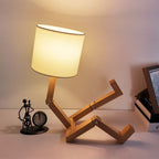 Clérachic wooden table lamp with jointed limbs raised in a reclining pose, its shade lit warmly beside a clock and framed picture.