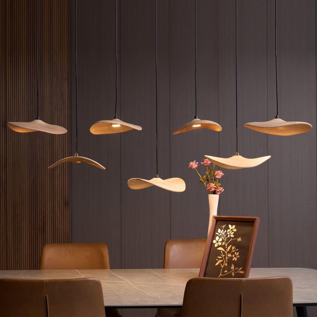 Lit Clérachic wooden leaf pendants over a stone dining table, with a vase of pink flowers and a framed print.