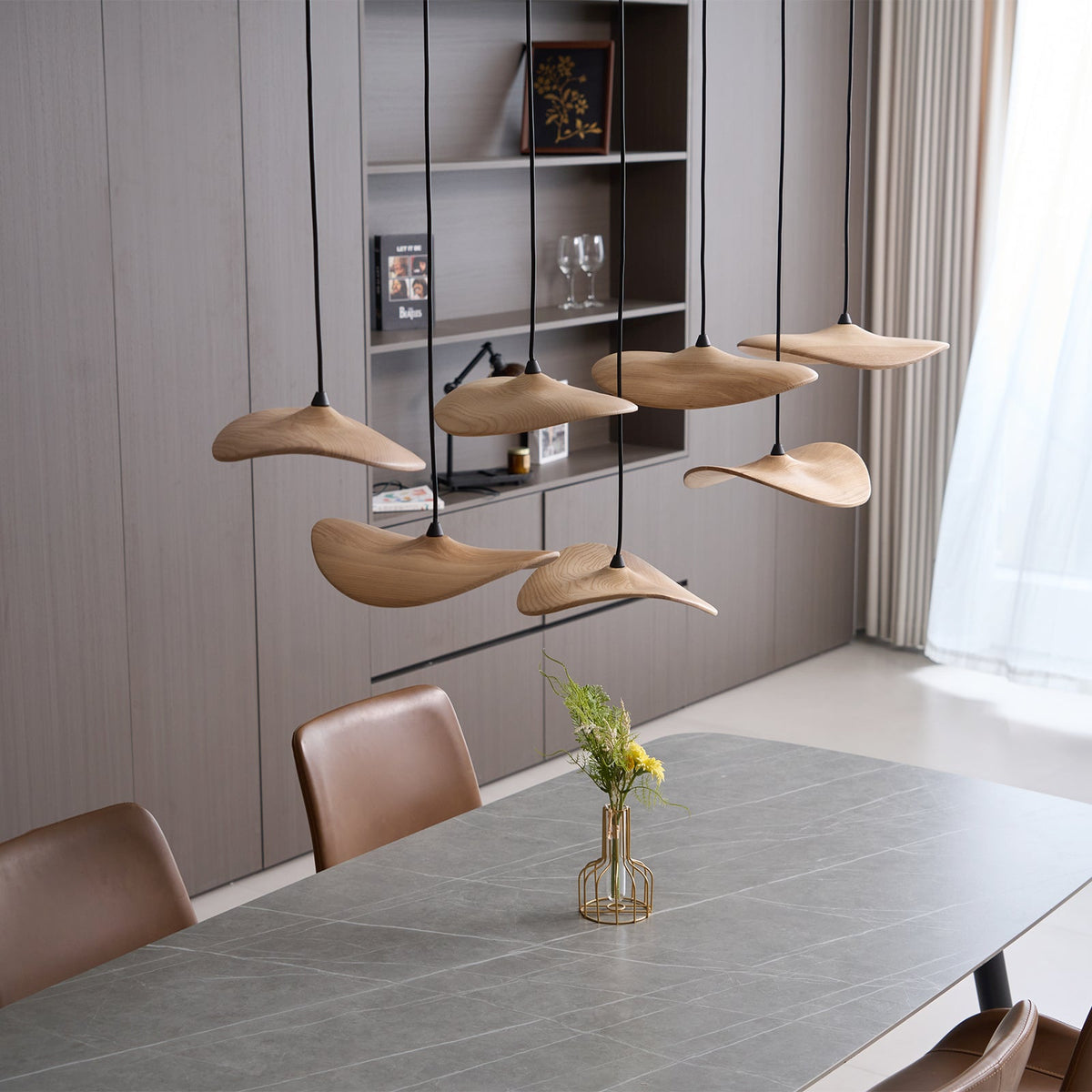 Clérachic wooden leaf pendants above a grey stone table, with a shelving unit and a vase of yellow flowers behind.