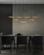 Seven Clérachic wooden leaf pendants over a dark dining area with black chairs and a charcoal wall panel.