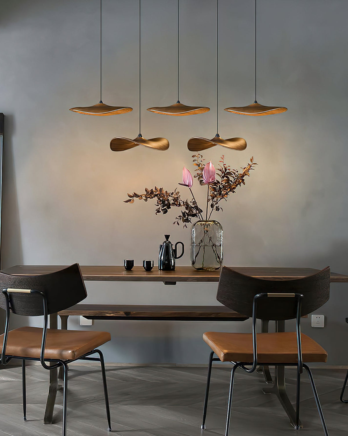 Five Clérachic leaf pendants lit over a dark dining table, with a vase of pink flowers and black chairs below.