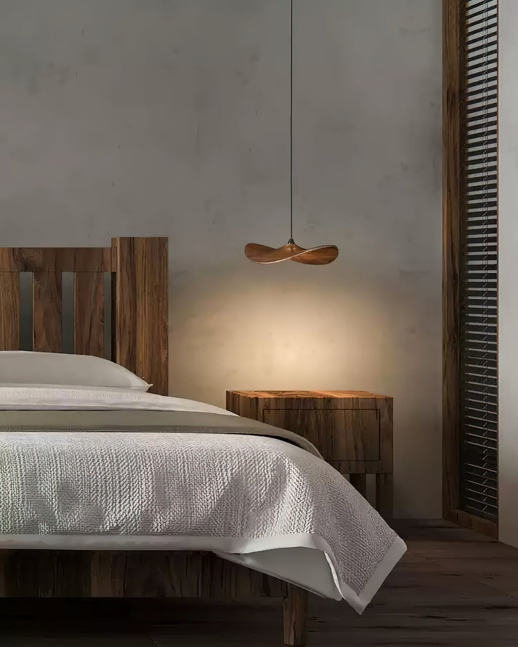 A single Clérachic wooden leaf pendant glowing beside a bed with white linen and a rustic wooden headboard.