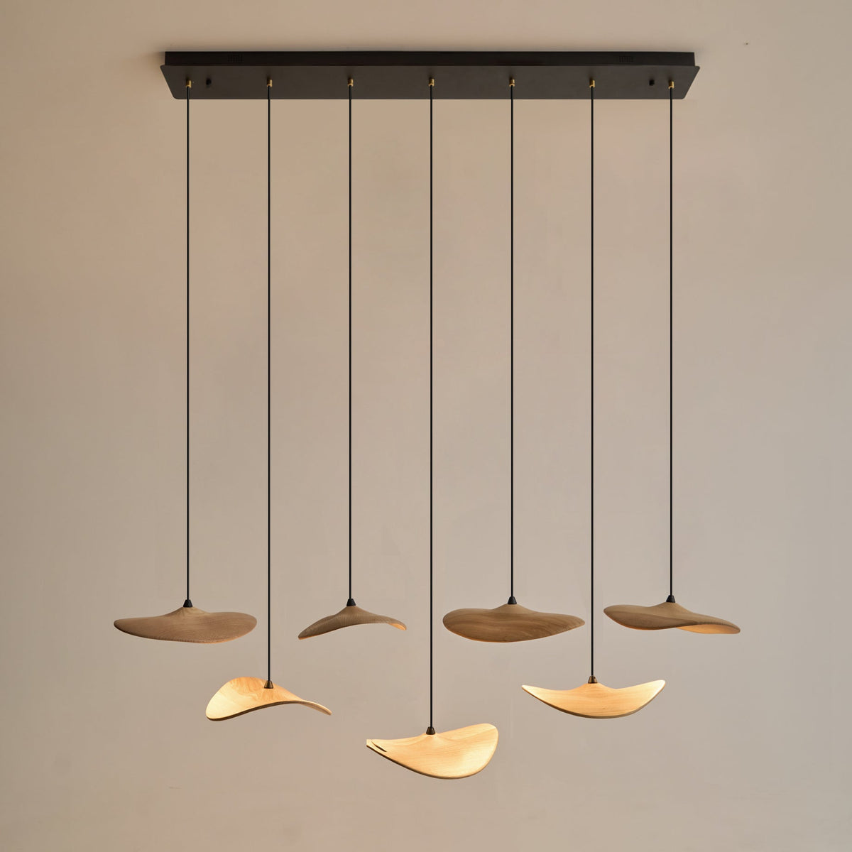 Seven Clérachic leaf pendants on a long black canopy, mixing dark and light wood, hung against a pale wall.