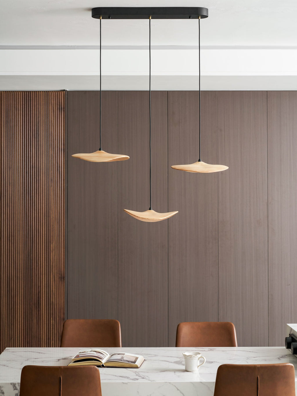 Three Clérachic leaf pendants over a marble island with an open book, a mug and leather chairs, before a ribbed wall.