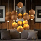 A Clérachic wood-sphere pendant glowing warm over a grey sofa, with framed monochrome prints and a dark timber wall behind.