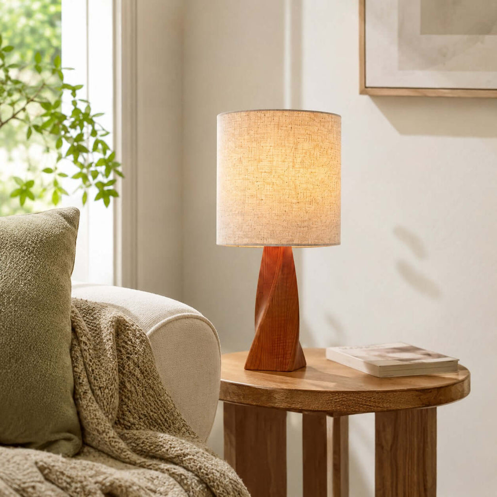 Soft light from this Clérachic table lamp glows through a linen drum shade on a round wooden side table beside a cream armchair and a green cushion.