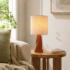 Soft light from this Clérachic table lamp glows through a linen drum shade on a round wooden side table beside a cream armchair and a green cushion.
