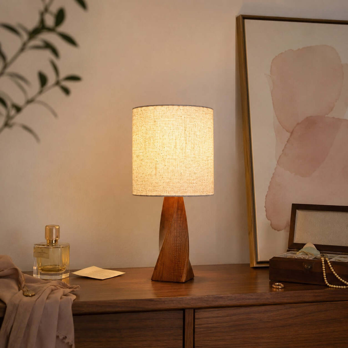 A twisted wood base and lit beige shade stand on a dark wooden dresser beside a perfume bottle, a keepsake box and a pink abstract painting, from Clérachic.