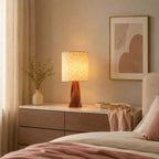The lit linen shade of this Clérachic table lamp glows on a pale chest of drawers beside a bed with rose bedding, a small bud vase and a soft print.
