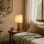 A bedside nook with a small round wooden table holds this Clérachic table lamp, a book and a mug, next to a cream daybed with a green throw.
