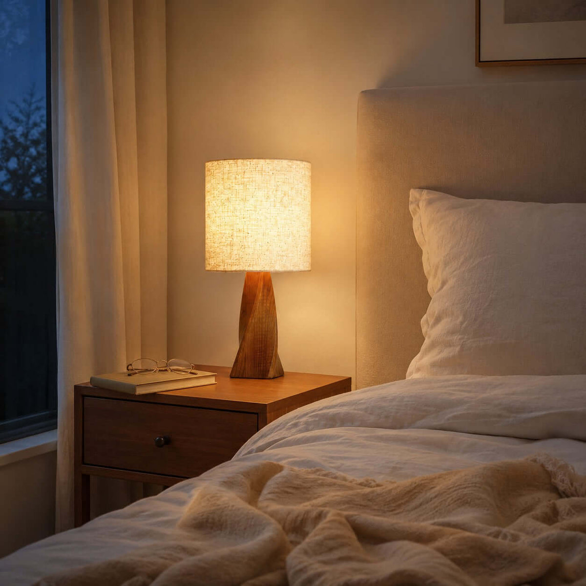 At dusk this Clérachic table lamp glows warm on a dark bedside table with a book and glasses, next to a bed with cream and peach bedding.