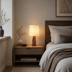 Beside a bed with grey linen and a taupe throw, this Clérachic table lamp lights a wooden nightstand with a small vase and a dark bowl.