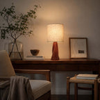A lit Clérachic table lamp stands on a dark wooden console with a glass vase of branches, a framed picture and a wooden bowl, beside a woven stool.