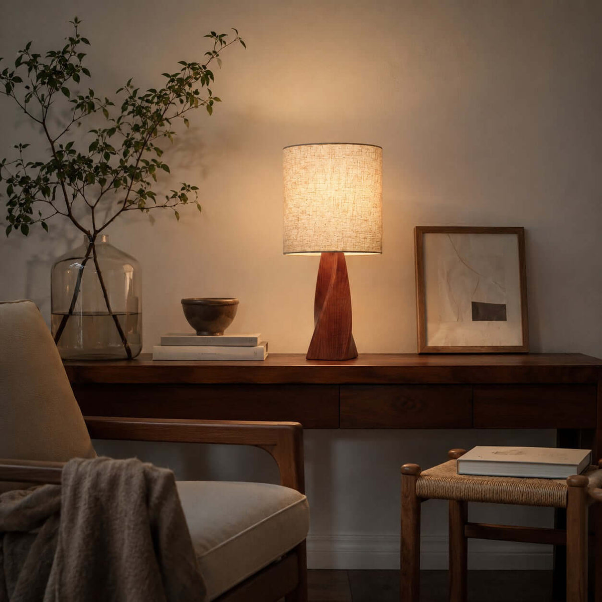A lit Clérachic table lamp stands on a dark wooden console with a glass vase of branches, a framed picture and a wooden bowl, beside a woven stool.