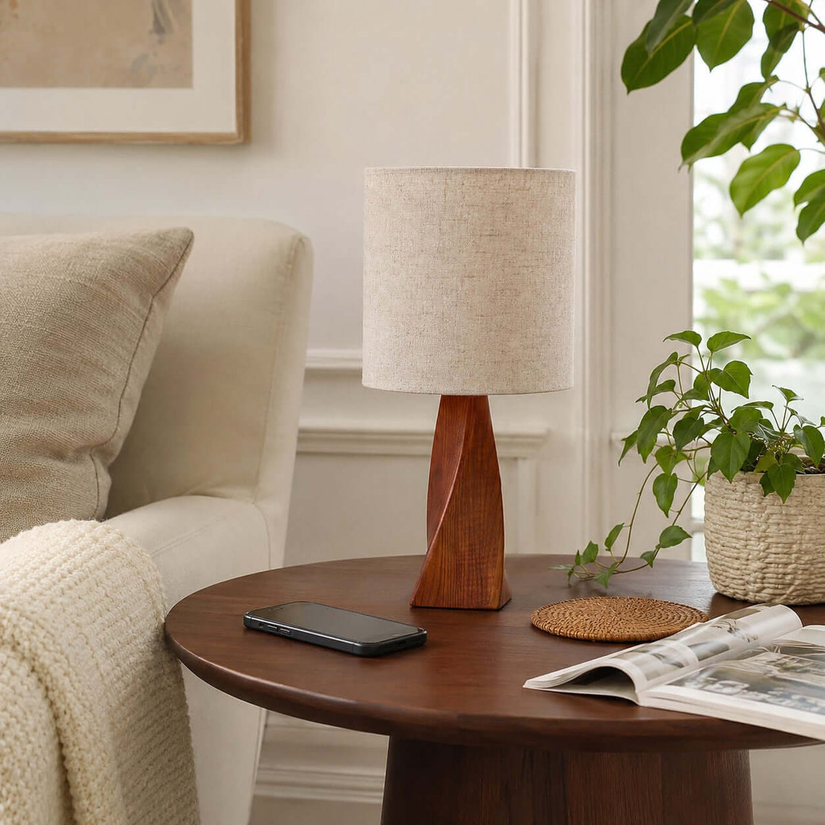 The twisted wooden base of this Clérachic table lamp sits on a round dark wooden table, with a phone, a coaster and a magazine beside a cream sofa.