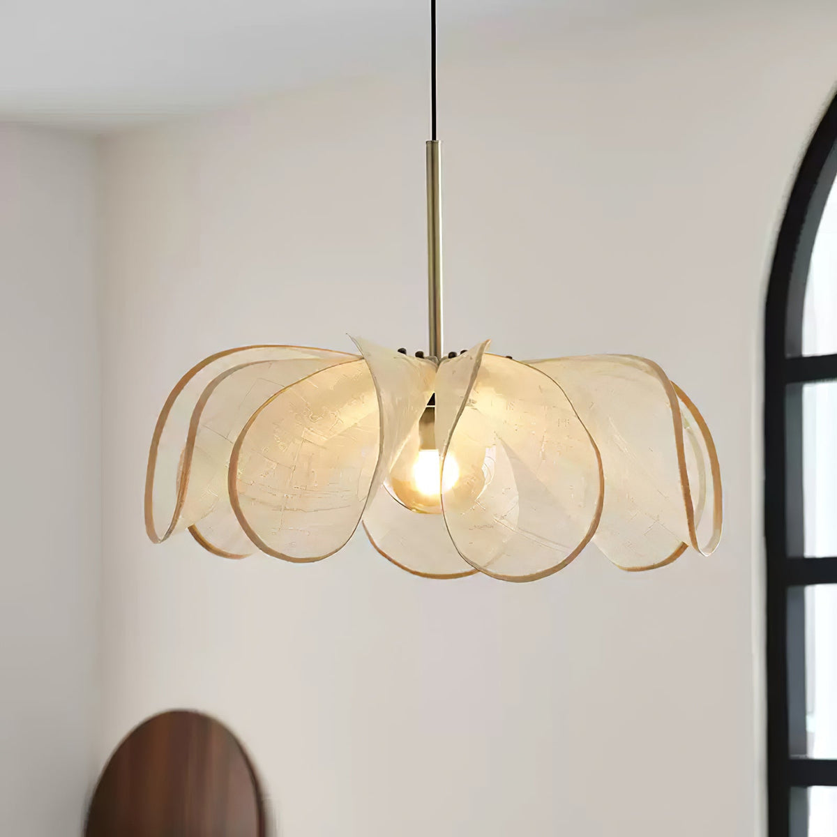 Lit abaca bloom shade with gold rim edging, seen against a pale wall with an arched window, a Clérachic pendant.