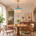 A Clérachic pendant with a blue and white woven drum shade hangs over a round wooden table with fan-back chairs and flowers.