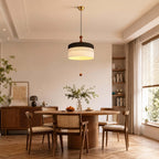 A black and white Clérachic pendant hangs in a wide dining room with a round table, rattan chairs and a bookshelf behind.