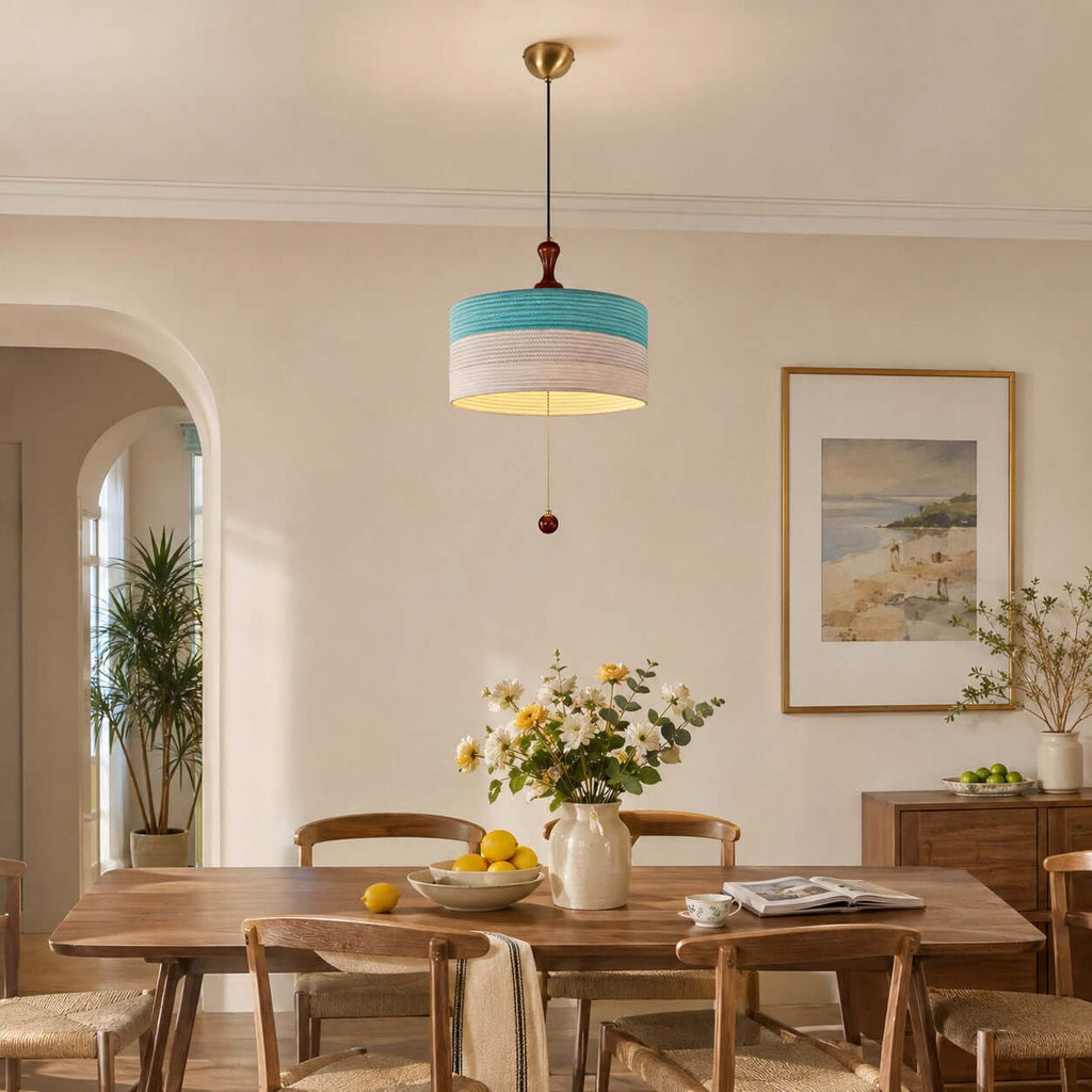 A blue and white Clérachic pendant hangs over a timber dining table with yellow flowers, lemons and a framed seaside print behind.