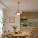 This Clérachic pendant in pink has a brass-coloured cap and a glowing globe, hung above a round wooden table with flowers.