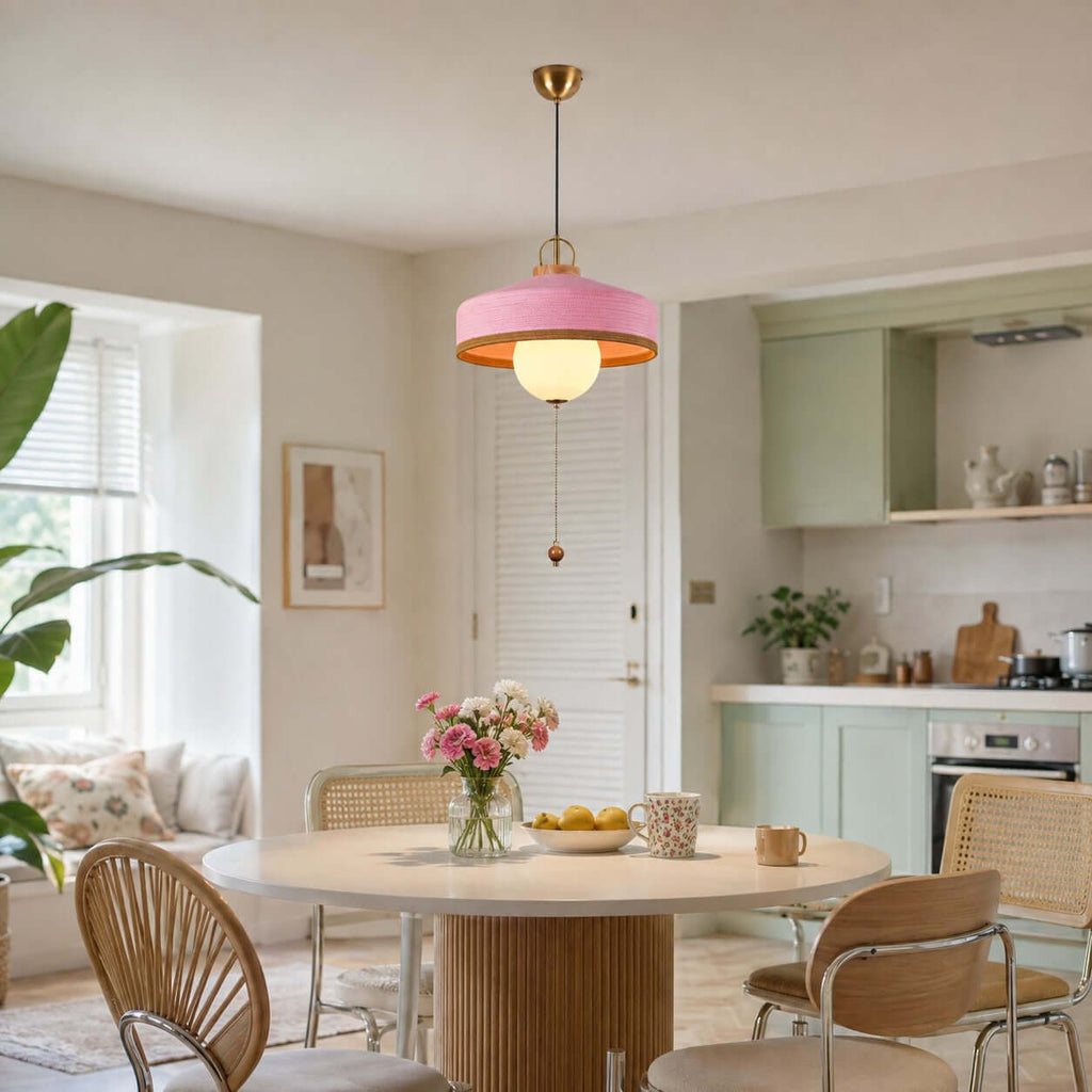 This Clérachic pendant with a pink woven shade hangs over a pale table with spring flowers, beside a sage-green kitchen.