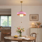 A pink Clérachic pendant with a glass globe hangs by a wall with a framed print, above a table with an open book and lemons.