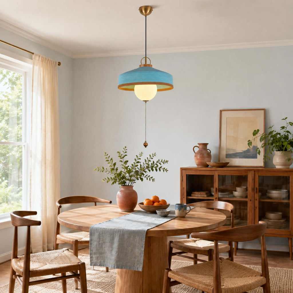 Blue woven shade and glowing globe on this Clérachic pendant, hanging in a dining room with a wooden table and tall curtains.