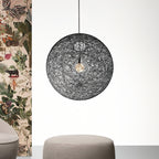 A black woven spherical Clérachic pendant light beside a panel of botanical wallpaper, above a grey round pouffe.