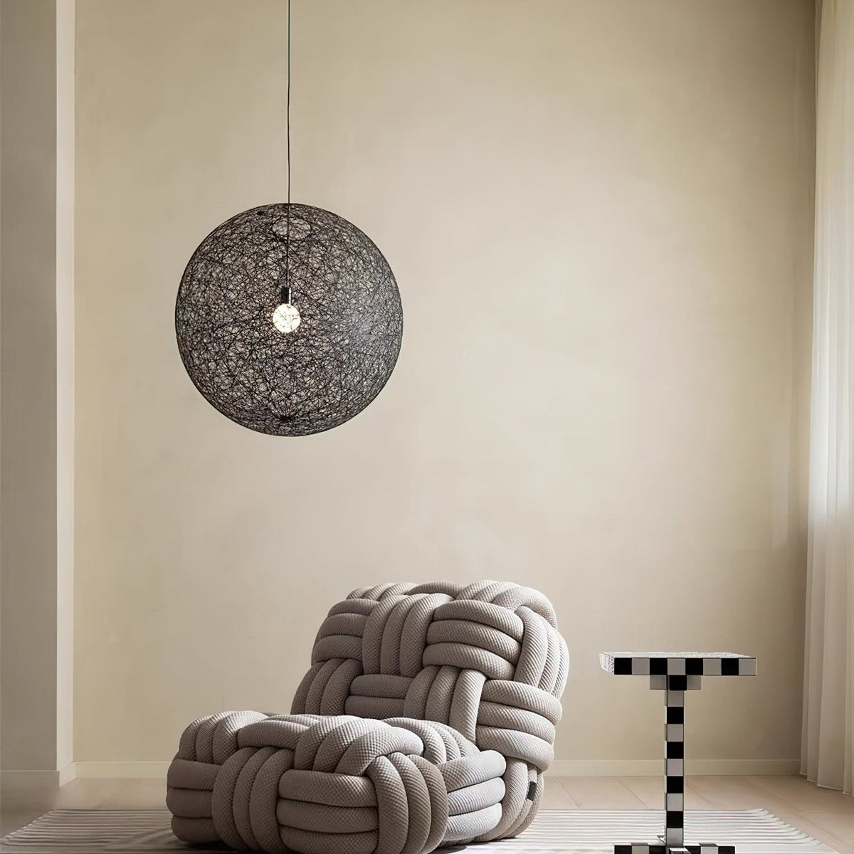 A black woven Clérachic pendant light hung in a calm beige room, above a knotted rope armchair and a chequered side table.