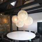A cluster of off-white woven Clérachic pendant lights lit above a round white table, under dark roof beams.