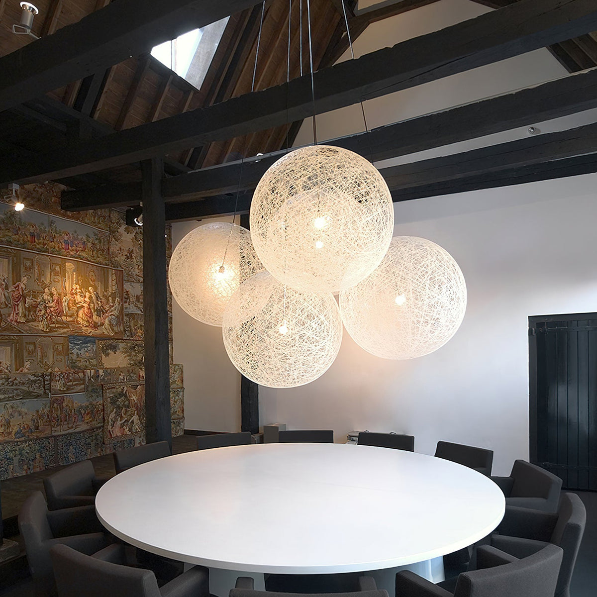 A cluster of off-white woven Clérachic pendant lights lit above a round white table, under dark roof beams.