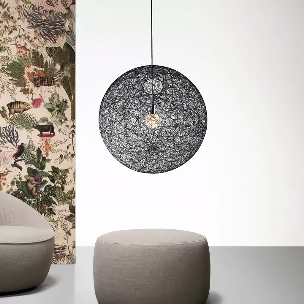A black woven spherical Clérachic pendant light with its bulb glowing, beside botanical wallpaper above a pouffe.