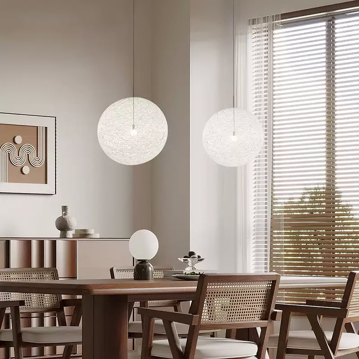 Two off-white woven Clérachic pendant lights above a wooden dining table with rattan chairs, in a room with a framed print.