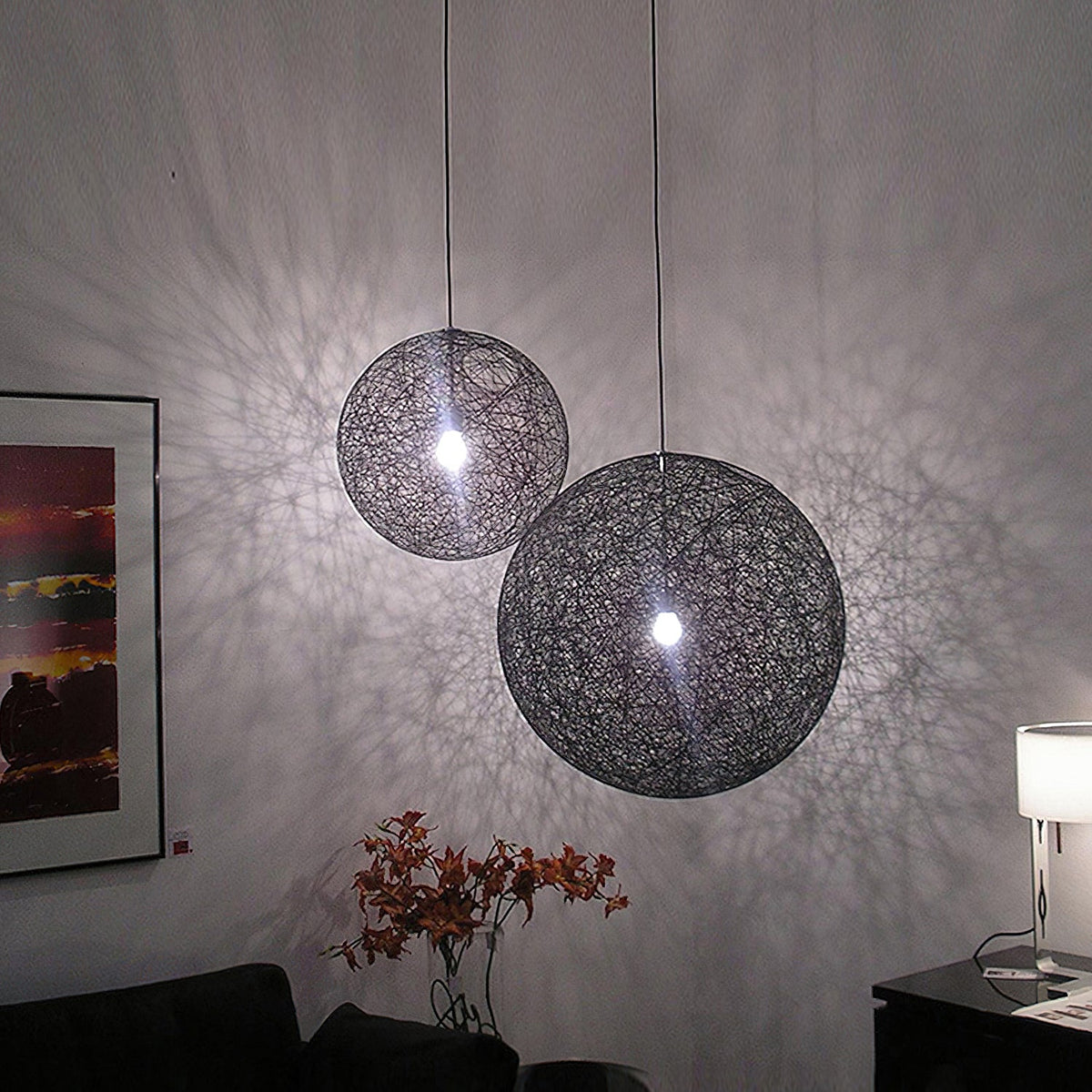 Two black woven Clérachic pendant lights of different sizes lit, throwing a lace-like shadow pattern across a grey wall.
