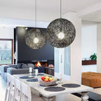 Two black woven Clérachic pendant lights lit above a pale dining table, with a dark fireplace wall and orange ottoman beyond.