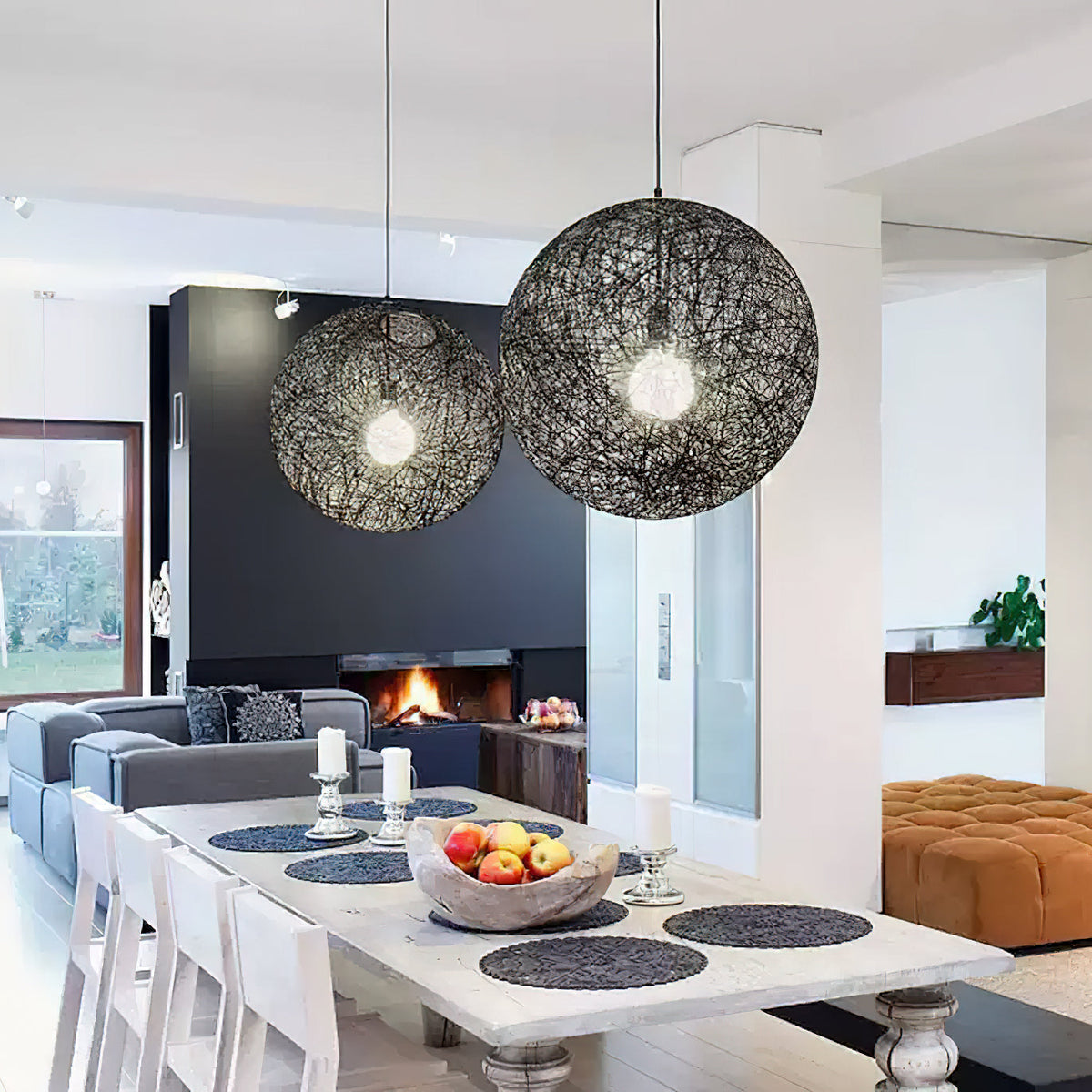 Two black woven Clérachic pendant lights lit above a pale dining table, with a dark fireplace wall and orange ottoman beyond.