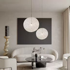 Two off-white woven Clérachic pendant lights in front of a dark panel, above a curved cream sofa and a marble coffee table.
