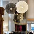 A black and an off-white woven Clérachic pendant light lit in a double-height room, with their reflections in a mirrored wall.