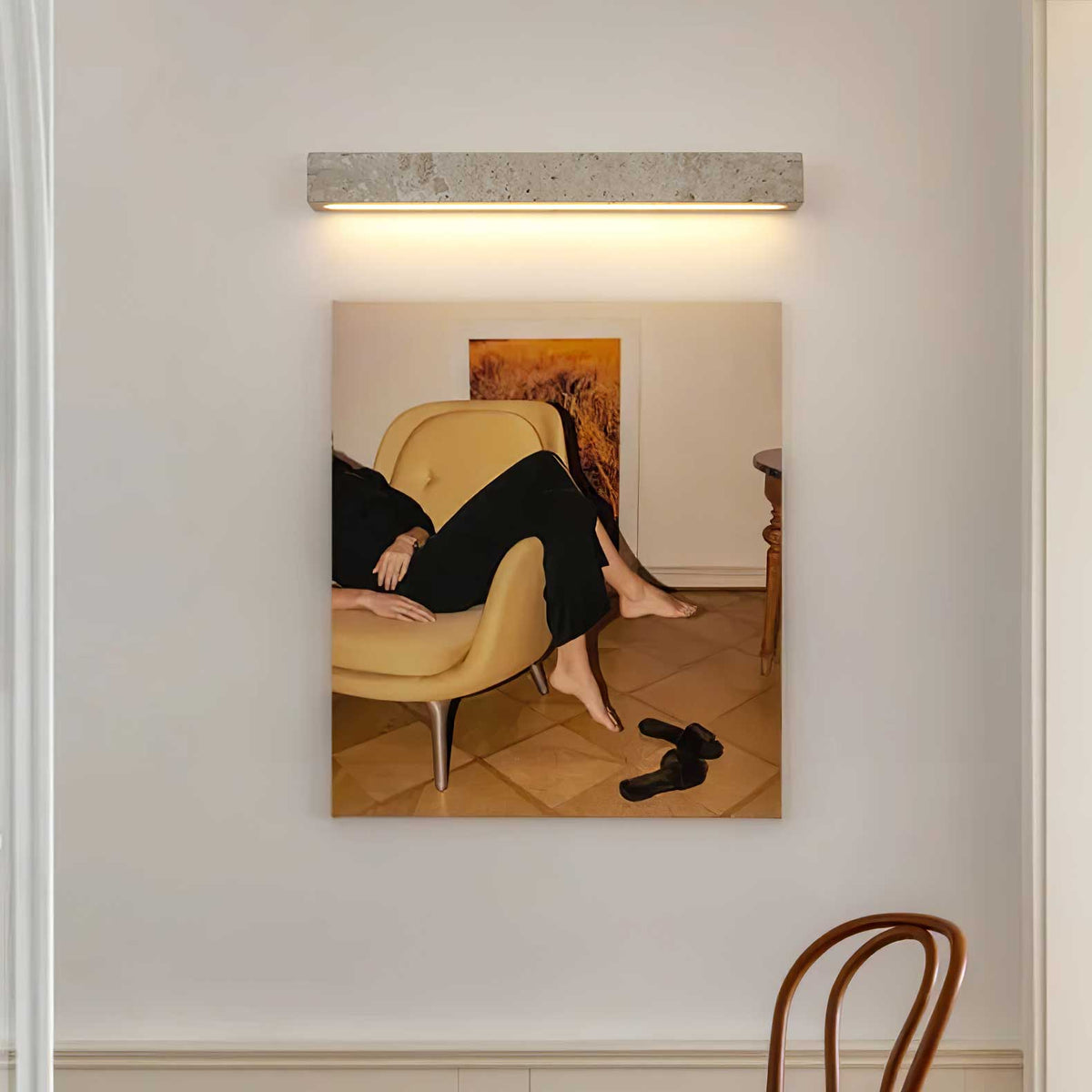 A horizontal Clérachic wall light in pale travertine casts a warm glow over a framed photograph of a figure in a yellow armchair.