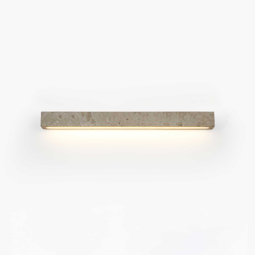 A slim pale travertine Clérachic wall light with its warm LED strip glowing below, shown on a white backdrop.