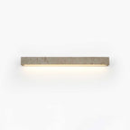 A slim pale travertine Clérachic wall light with its warm LED strip glowing below, shown on a white backdrop.