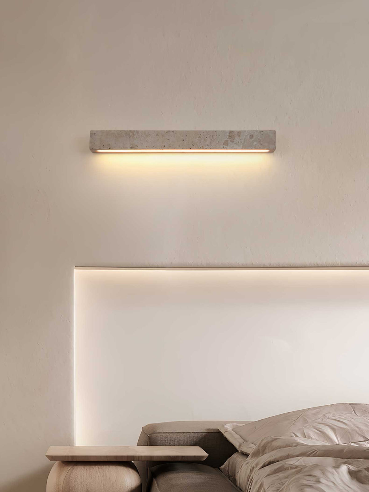 A Clérachic travertine wall light glows warm on a cream wall above a bed, with a lit recess beneath it.