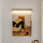 A pale travertine Clérachic wall light hangs above a framed photograph of a yellow armchair, a wooden chair at the corner.
