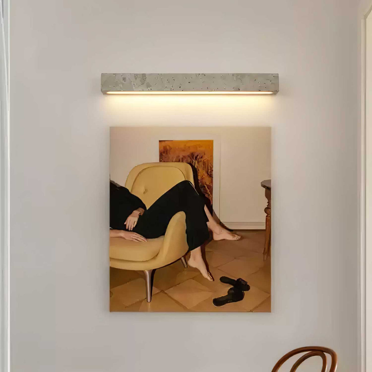 A pale travertine Clérachic wall light hangs above a framed photograph of a yellow armchair, a wooden chair at the corner.